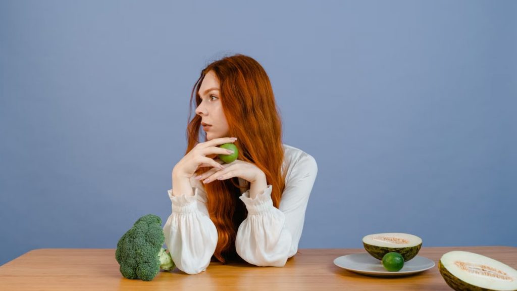 Thoughtful woman with broccoli, melon, and lime on a table.