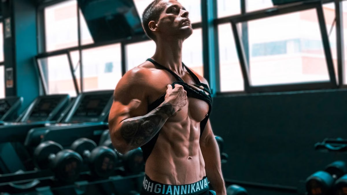Muscular man showing abs and tattoos in a gym setting.
